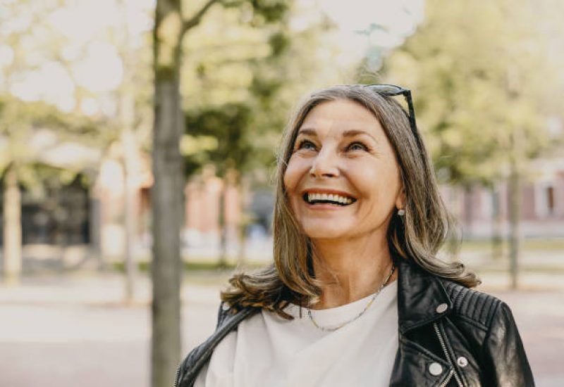 Cheerful overjoyed middle aged woman wearing sunglasses on her head and black leather jacket over white blouse enjoying peaceful beautiful morning while waking in park, looking up with broad smile